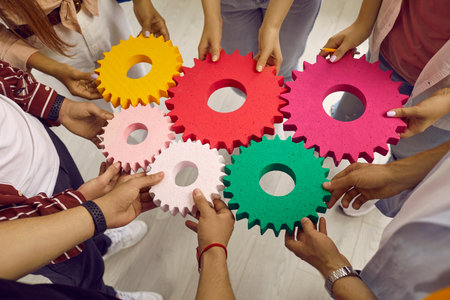 Creative young people hold colored gears of different sizes symbolizing coordinated teamwork. Top view from angle of hands of men and women standing in circle. Concept of team and teamwork.の写真素材