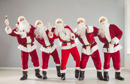 Full length portrait of happy funny people in Santa Claus red costumes standing in a row holding clock showing five minutes to midnight. New Year eve and Christmas holidays celebration.の写真素材