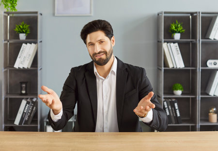Puzzled uncertain man ceo dressed in formal black suit, shrugging shoulders as doesnt know how to solve problem, posing at table in office room, faces some difficulties. Confused businessmanの写真素材