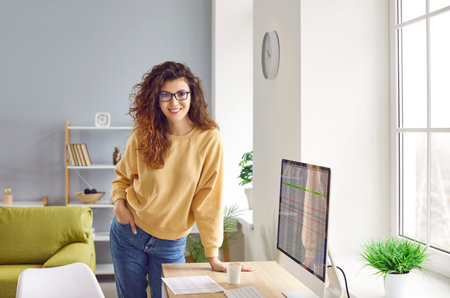 Portrait of happy woman who works at home. Young girl with beautiful long hair in casual yellow sweatshirt, jeans and eyeglasses standing by desk with computer and coffee looking at camera and smilingの写真素材