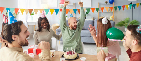 Joyful man raising hands while celebrating birthday with friends. Group of happy diverse people at desk wearing party hats cheering, clapping, enjoying cake and gifts in decorated living room. Banner.の写真素材