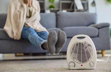 Man with blanket near heater at home. Sitting on a sofa in a cold room, he relies on a portable...