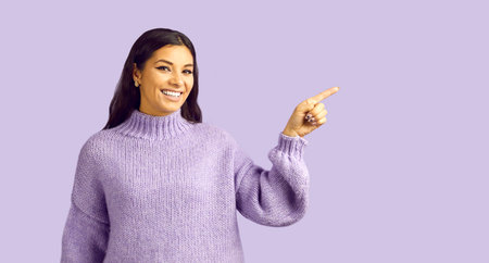 Happy fashion model in light purple knit woolen sweater shows something on lavender background. Beautiful young brunette woman wearing warm purple wool jumper points to side at blank text copy spaceの写真素材