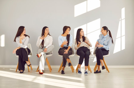 Young happy smiling female business people sitting in a row on chairs discussing work project in empty meeting room. Company employees women talking about new startups. Team work conceptの写真素材