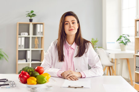 Portrait of cheerful young friendly female doctor nutritionist in medical uniform sitting on workplace in office. Smiling woman dietician looking at camera. Healthy nutrition and diet concept.の写真素材