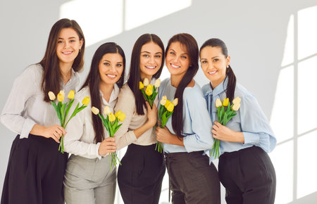 Group portrait of young female business colleagues with flowers on International Womens Day. Team of happy beautiful ladies with yellow tulips celebrating holiday together in officeの写真素材