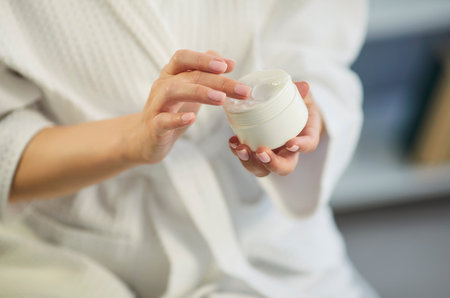 Closeup of woman holding jar with moisturizing cream in hands touching it with finger going to apply on body for healthy skin. Female person in white bathrobe doing beauty procedure. Skin care.の写真素材