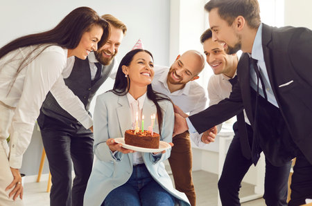 Team of business people celebrating a colleagues birthday at work. Group of coworkers and friends giving a cake with candles to a happy, smiling young woman in a party capの写真素材