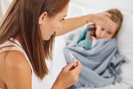 Worried mother holding thermometer, measuring temperature of sick child lying in bed wrapped in plaid. Caring woman sitting on bed, feeling forehead of little ill kid suffering from fever, cold or fluの写真素材