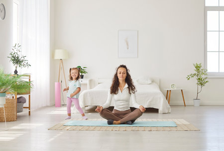Happy calm mother resting in lotus pose, breathing techniques, mindfulness practice with little daughter, sporty friendly sitting, doing mind, body meditation, fitness classes to improve life balanceの写真素材