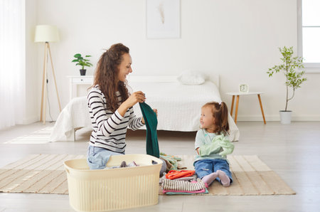 Mother and child work together folding clean clothes from a laundry basket at home. Shared housework moment highlights family togetherness, organization, and care in a warm, household setting.の写真素材