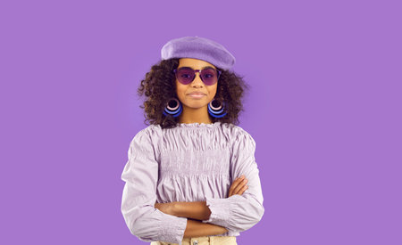 Child in modern outfit posing in fashion studio. Beautiful kid in trendy clothes isolated on purple background. Portrait of pretty African girl in lilac top, purple beret hat and sunglassesの写真素材