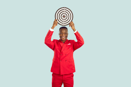 Smiling African American man in stylish red suit who is holding target over his head on pastel light...