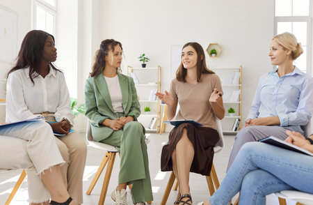 Women group meeting in a training circle. Colleagues share ideas and support each other in a bright office, seated in chairs. Collaboration and wellbeing are encouraged. Team growth concept.の写真素材