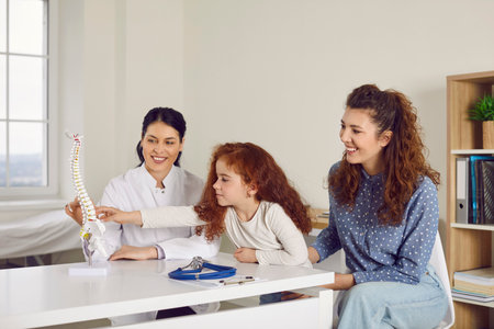 Curious kid together with mum visits doctor and learns about human skeleton. Happy woman and her daughter looking at orthopedic spine model on desk in pediatricians office. Children, medicine conceptの写真素材