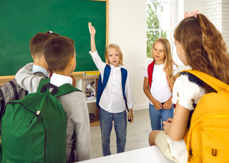 Little schoolboy wants to say something and raises his hand while talking to his classmates. Schoolchildren with colorful backpacks on their shoulders are about to go home after school lessons.の写真素材