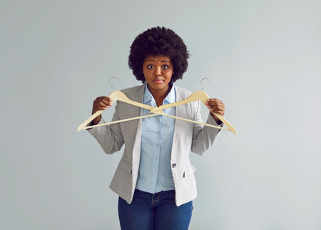 Woman has nothing to wear or cant decide on outfit. Portrait of beautiful Afro American model in suit jacket with funny confused face expression holding empty wooden clothes hangers. Fashion conceptの写真素材