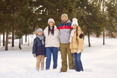 Full length portrait of happy smiling family of four with two kids walking in winter park and looking at camera. Young parents and children enjoying time together outdoors in snowy forest.の写真素材