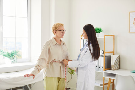 Young woman doctor cardiologist or physiotherapist in lab coat measuring waist of female patient with tape measure during regular medical physical examination in clinic. Weight loss and diet concept.の写真素材