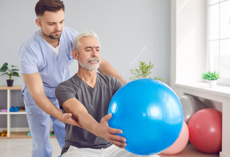 Focused doctor or nurse assists an elderly patient during a rehabilitation session with fitball...