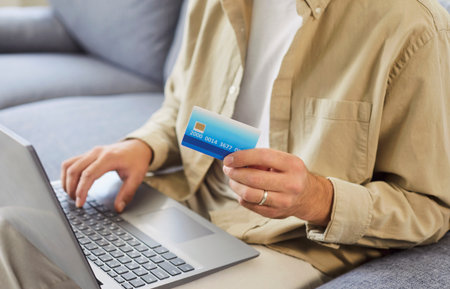 Man uses laptop, credit card for online payment. He verifies banking details and enters checkout data on the sofa at home, focusing on ecommerce shopping and finance. Internet banking concept.の写真素材