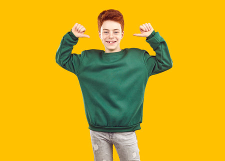Achieved it myself. Happy proud satisfied self assured male child youth model leader boy in modern mockup shirt points thumbs at himself and looks at camera on yellow studio backgroundの写真素材