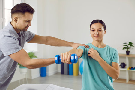 Happy young girl doing exercises with dumbbells at modern physiotherapy center. Male physiotherapist...