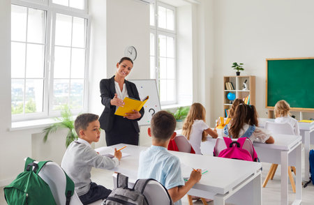 Cheerful female teacher teaching kids in classroom at school. View from behind of cute little boys...