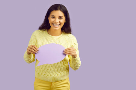 Smiling millennial Latin girl isolated on purple studio background hold speech bubble show message...