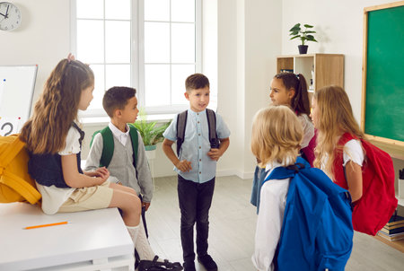 Group of friendly elementary school students are talking in classroom after school lessons. Boys and girls 6-7 years old wear colorful backpacks on their shoulders and communicate in school classroom.の写真素材