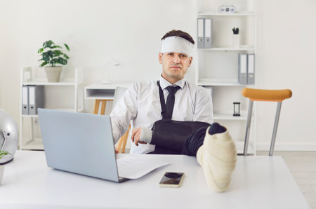 Sad businessman with a broken arm and leg, injured from a trauma or accident, continues working in the office. His negative mood showing perseverance in a challenging business environment.の写真素材