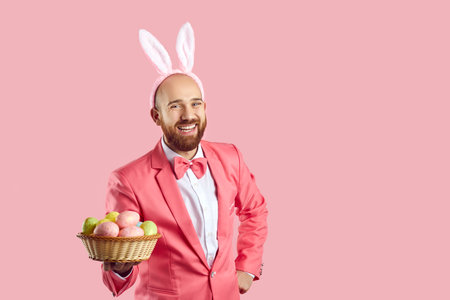 Studio shot of happy bald young man with ginger beard, wearing pink suit, bow tie and funny bunny ears standing on pink background, looking at camera, holding wicker bowl of Easter eggs and laughingの写真素材