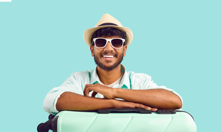 Happily excited Indian male tourist with suitcase waiting for trip abroad for weekend. Handsome bearded man in summer hat and sunglasses leans on suitcase and smiles on light blue background. Banner.の写真素材