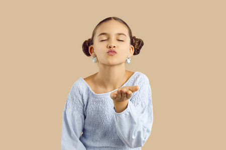 Funny kid blows air kiss. Cute little child wishes you love on Valentines Day and sends kisses. Studio shot of pretty girl with hair buns and stylish earrings closing her eyes and pouting her lipsの写真素材