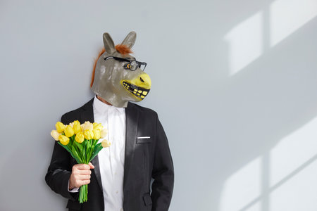 Unidentifiable man in rubber donkey head mask holding bouquet of flowers. Male in formal suit, white shirt and glasses standing at grey wall with yellow tulip flowersの写真素材