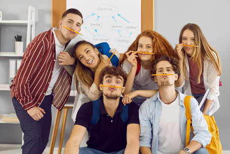 Portrait of funny student friends having fun and fooling around putting pencils between nose and lips. Cheerful young people holding pencils as mustaches posing for camera in classroom.の写真素材