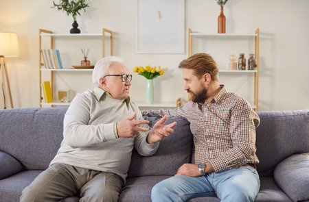 Young son and elderly father having conversation. White haired old man in his 60s or 70s sitting on sofa at home together with adult son, talking about life and sharing advice. Senior parents conceptの写真素材