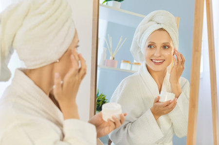 Young woman applying moisturizing cream on face in bathroom and looking at mirror. Female person in white bathrobe doing morning beauty procedure holding cosmetic jar. Body health and skin care.の写真素材