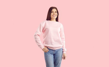 Woman in casual outfit posing in fashion studio. Portrait of happy attractive young European girl wearing plain pastel pink sweatshirt and blue jeans standing isolated on solid pink colour backgroundの写真素材