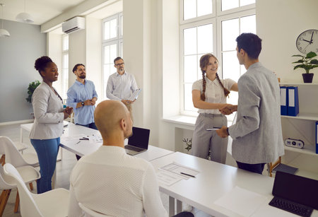 Business people man and woman greeting each other shaking hands and collegues looking at them in office. Corporative business meeting at working day. International corporation, team building concept.の写真素材