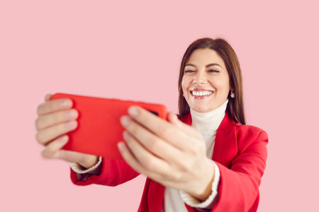 Happy pretty woman smiles and takes selfie on mobile phone isolated on pink background. Young girl in smart casual jacket and turtleneck with cheerful face expression takes selfie on modern smartphoneの写真素材