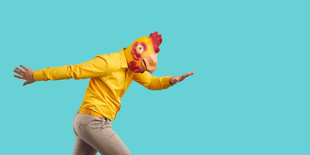 Cheerful funny man in strange rubber mask of chicken dancing isolated on turquoise background. Young man in yellow shirt, pants and with roosters head having fun near copy space. Banner.の写真素材