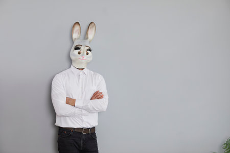 Businessman in a funny animal mask on a grey copy space background. Portrait of a business man wearing a white shirt and a rabbit mask standing with crossed arms by an empty grey studio wallの写真素材