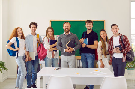 Group portrait of friendly male teacher and his college or high school students. Smiling teacher and students with backpacks on shoulders stand in row on background of blackboard and smile at camera.の写真素材