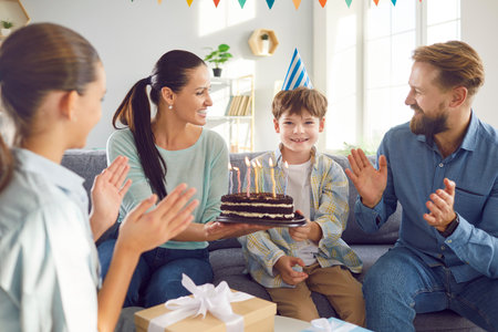 Happy birthday family at home, child boy ready blowing candles on birthday cake, enjoys surprise, wearing party hat, parents and sister glad to congratulate, best wishes warm friendly relationsの写真素材