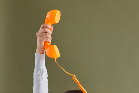 Human hand holding bright vivid orange landline phone receiver. Cropped shot of young lady raising up retro telephone receiver against khaki green blank copy space background. Customer support conceptの写真素材