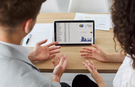 Company employees sitting back at the desk working in office looking at tablet screen with diagram...