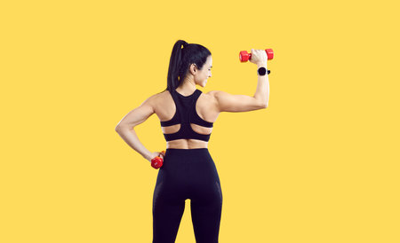 Young woman demonstrates muscles posing with back to camera with dumbbells in hands doing fitness to...