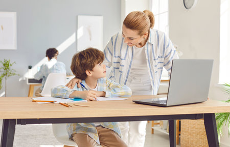 Happy Caucasian mother is helping young son with homework during online education. They are learning together using a laptop and internet, emphasizing family support and academic success.の写真素材