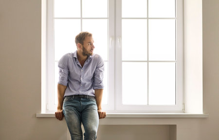 Man by window thinking about depression, anxiety. Soft daylight traces his pensive posture on the sill. He faces stress, solitude, and a health problem. Core concept: mental health struggle.の写真素材